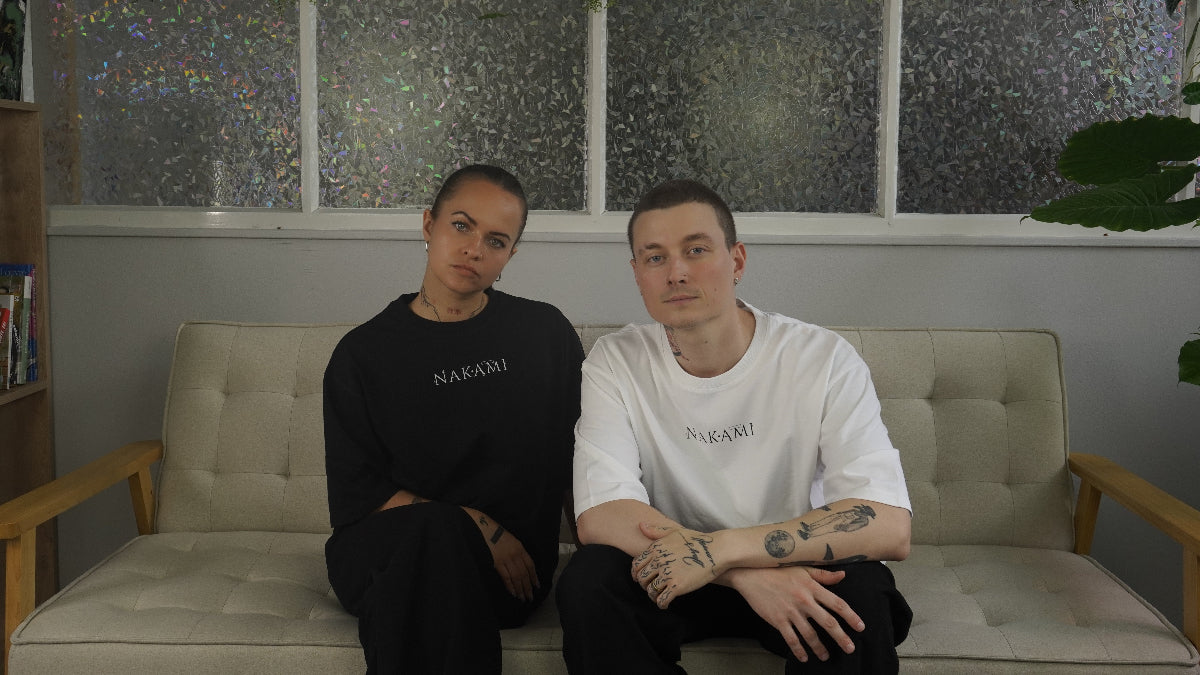 Ein tätowierter Mann und eine tätowierte Frau sitzen auf einer beigen Couch und schauen in die Kamera. Währenddessen tragen sie Anime Streetwear